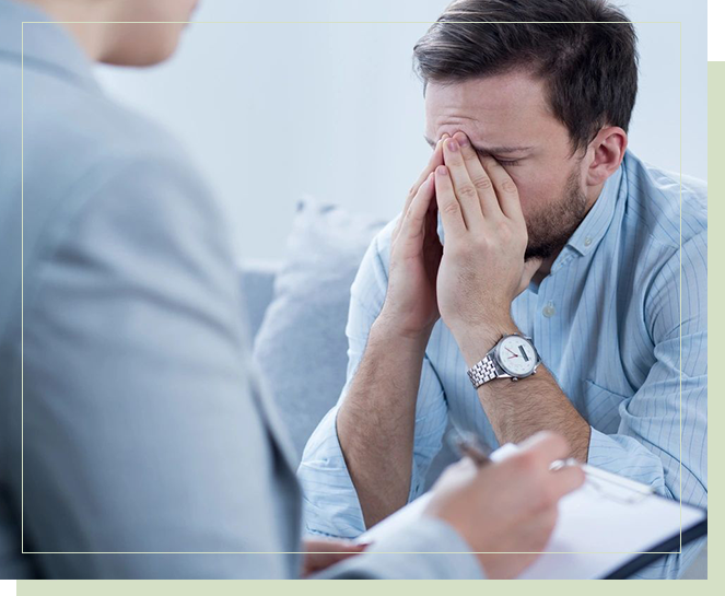 A man with his hands over his face as another person sits in front of him.